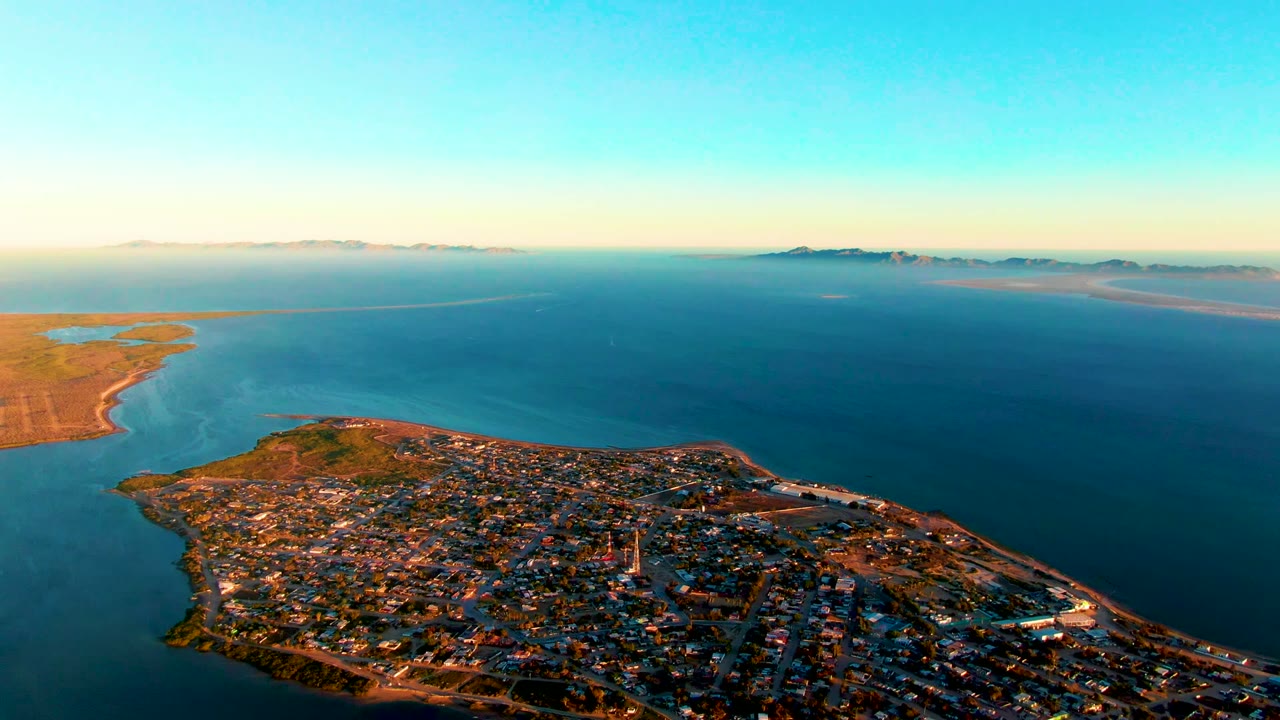 San Carlos appears from above with dense buildings along the lower edge, turquoise shallows at left, darker blue open water at center, thin horizon land, and no visible people.