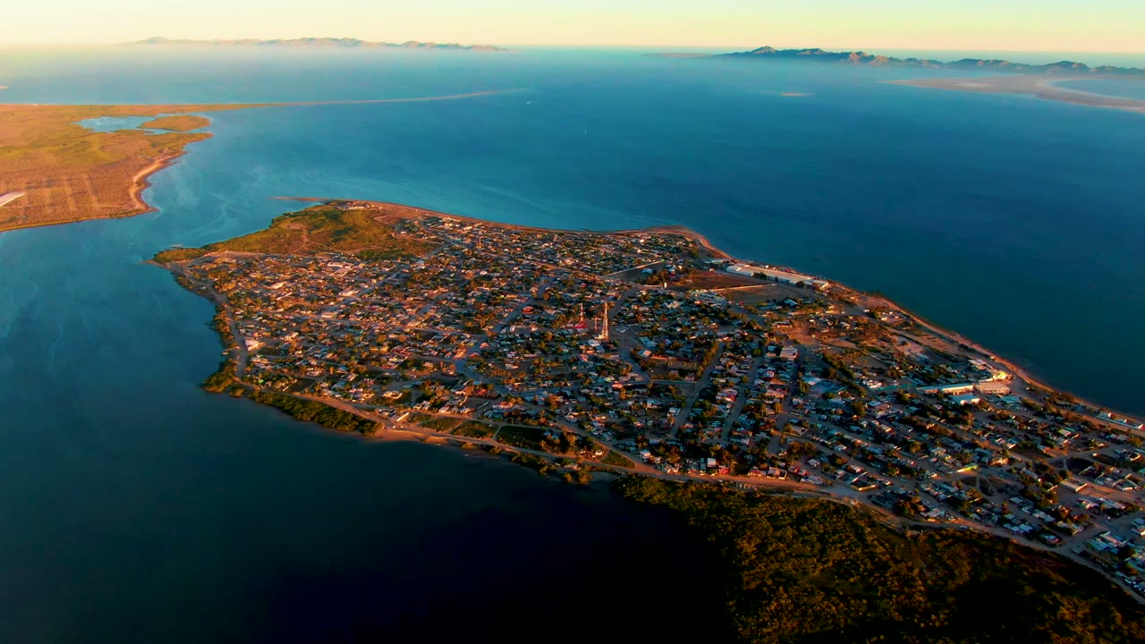 Opening aerial frame over San Carlos, Baja California Sur, showing the town on a peninsula with blue water around it, distant hills on the horizon, and no visible people.