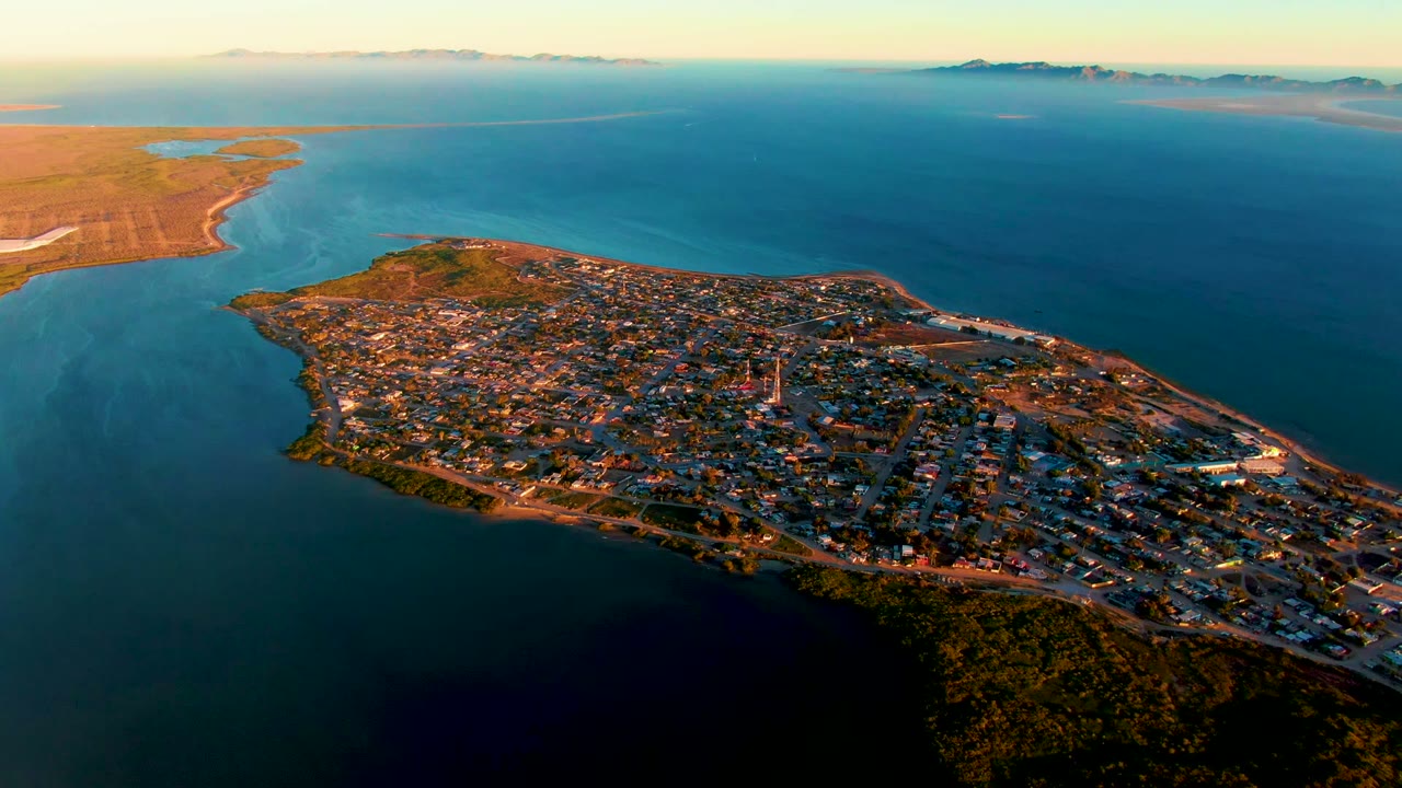 High aerial view over San Carlos, Baja California Sur, showing a town-filled peninsula, surrounding blue water, distant coastline, warm sunset light, and no visible people.
