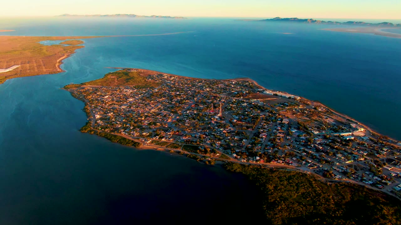 Coastal peninsula town seen from above with blue ocean and warm evening light.