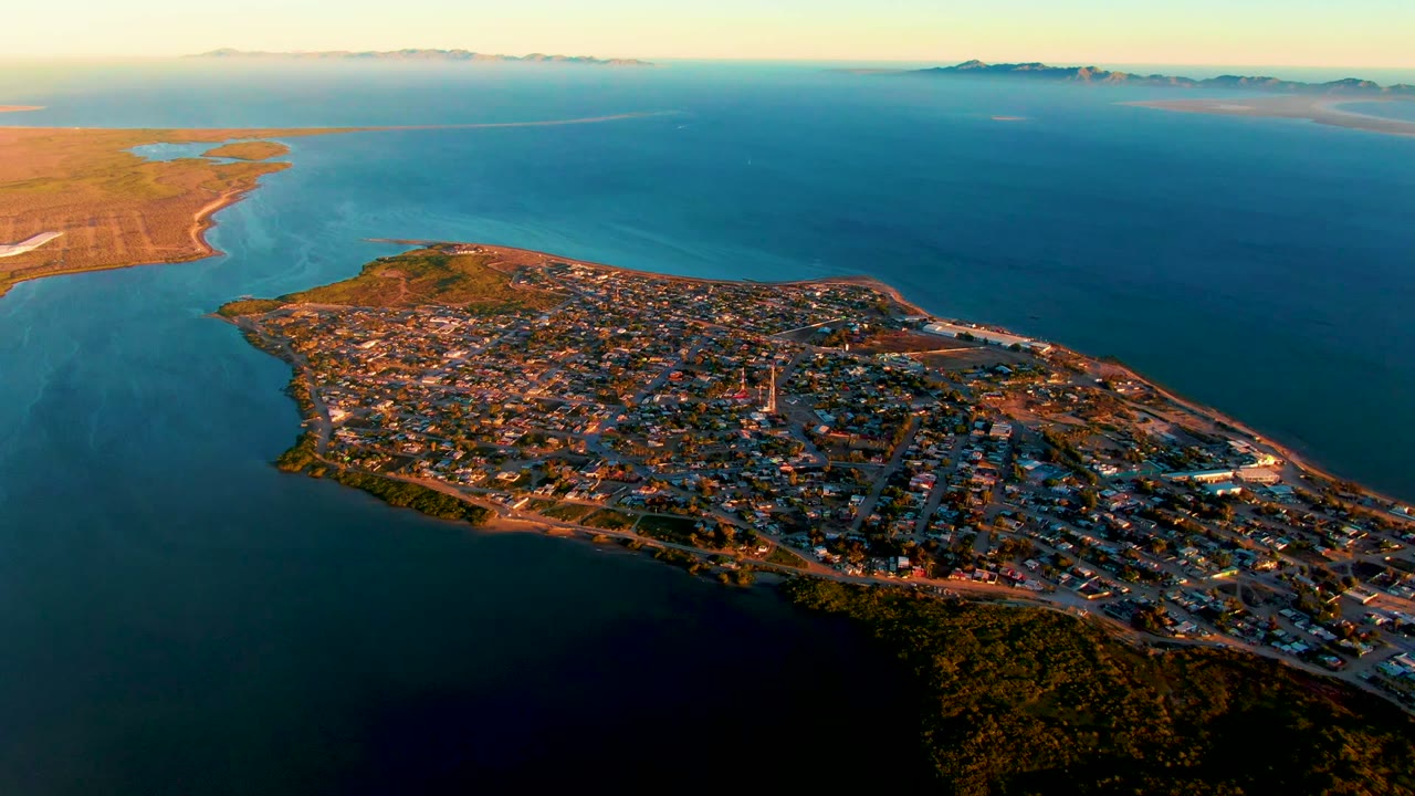 Wide aerial view of San Carlos, Baja California Sur, with the peninsula town extending across the frame, darker water in the foreground, open sea behind it, and no visible people.