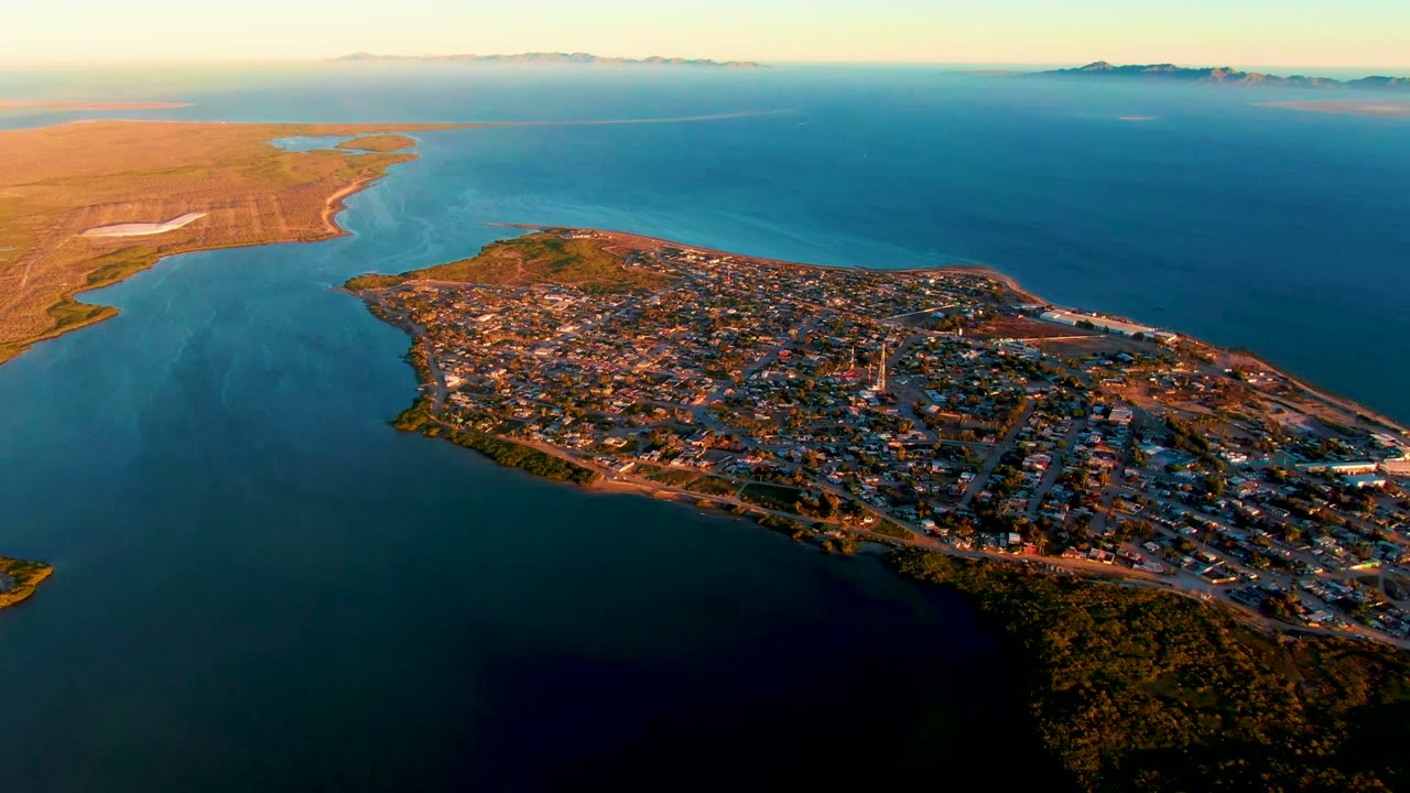 Opening aerial frame over San Carlos, Baja California Sur, with the town covering a peninsula, blue water on three sides, brown flats at left, distant mountains on the horizon, and no visible people.