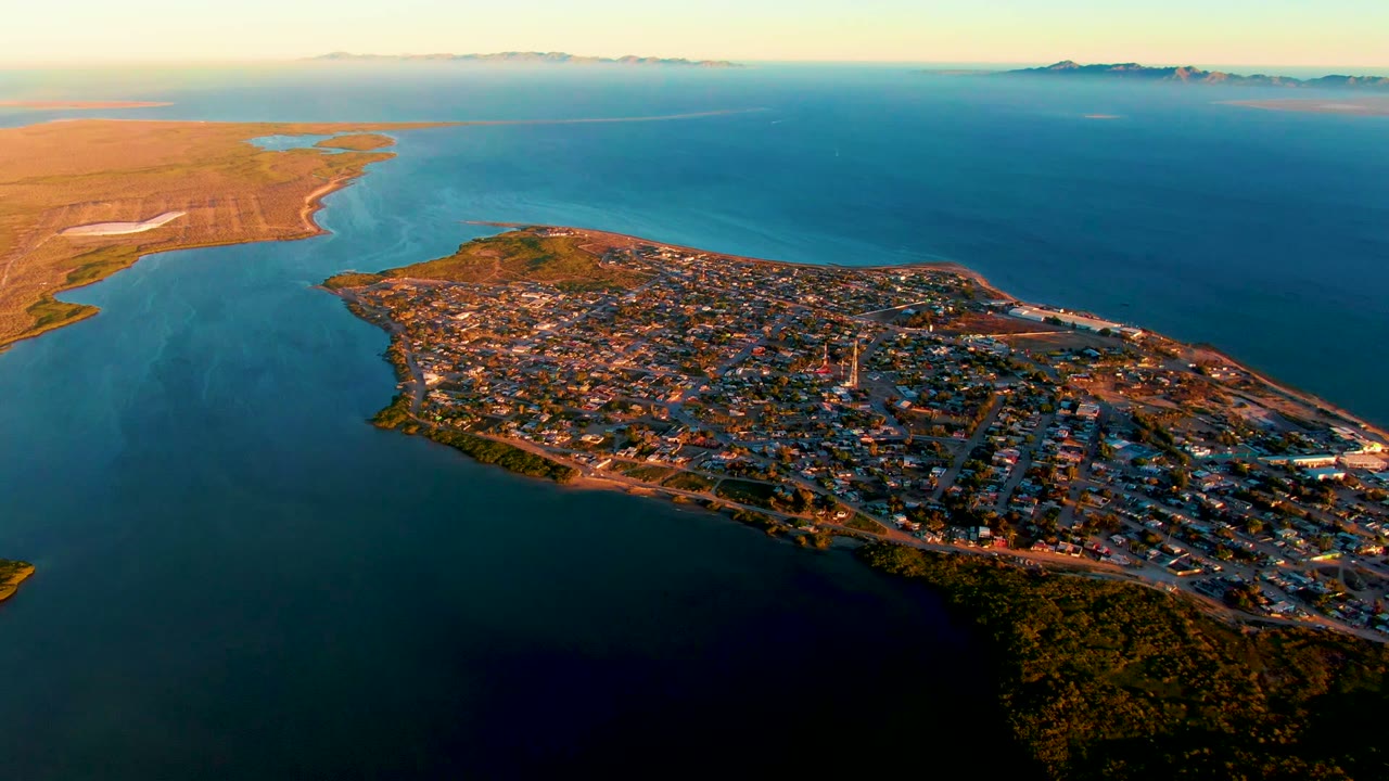 Aerial view of San Carlos with the peninsula centered lower in frame, dense buildings and roads visible, open blue water behind it, brown land across the channel, and no visible people.