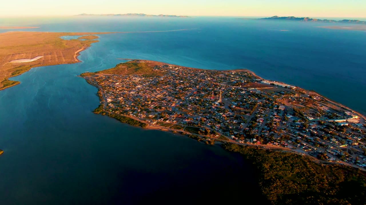 San Carlos appears from above with warm light across the roofs, a bright shoreline tracing the peninsula edge, deep blue water to the right, lighter channel water at left, and no visible people.