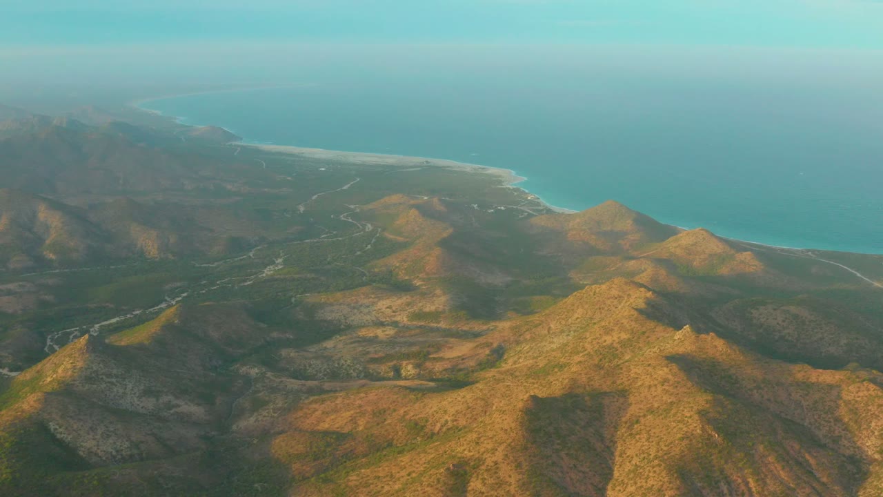 Opening aerial frame over Cabo Pulmo, Baja California Sur, with sunlit brown ridges in the foreground, green patches in the valleys, the blue sea beyond, and no visible people.