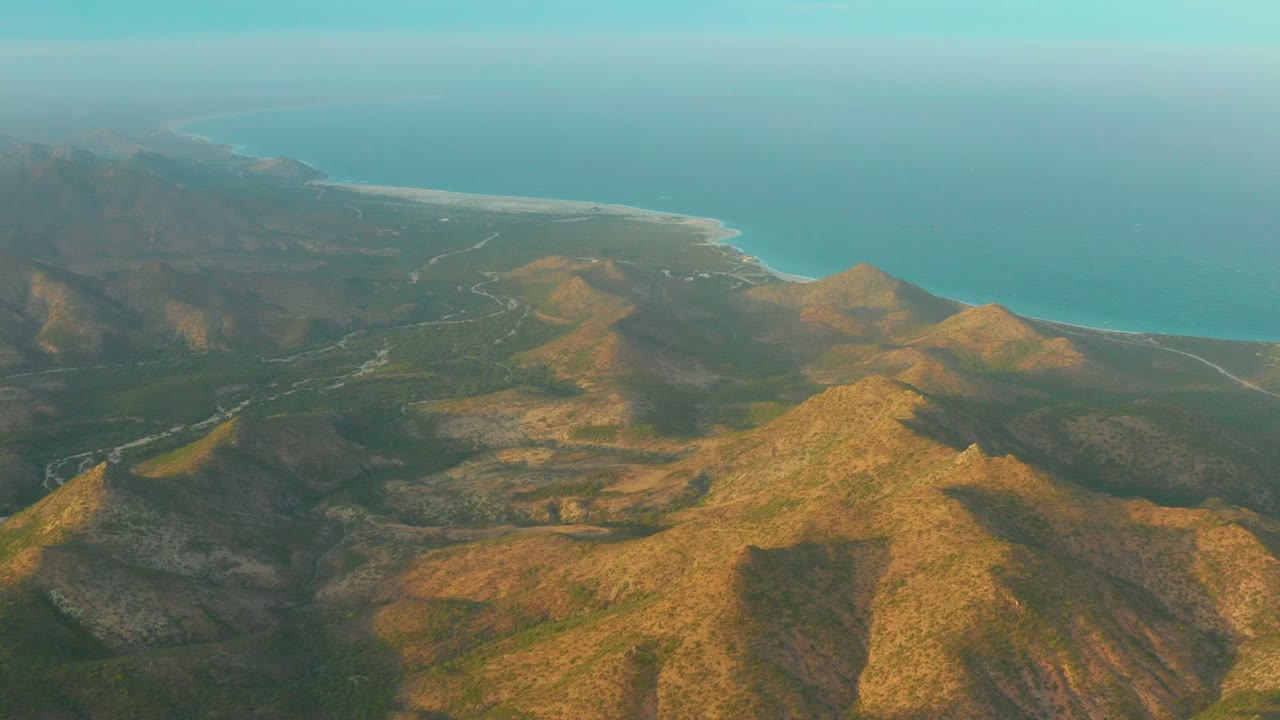 A later aerial frame shows the sunlit foreground ridge occupying more of the lower frame, with darker hills, pale road lines, blue ocean, and no visible people in Cabo Pulmo.
