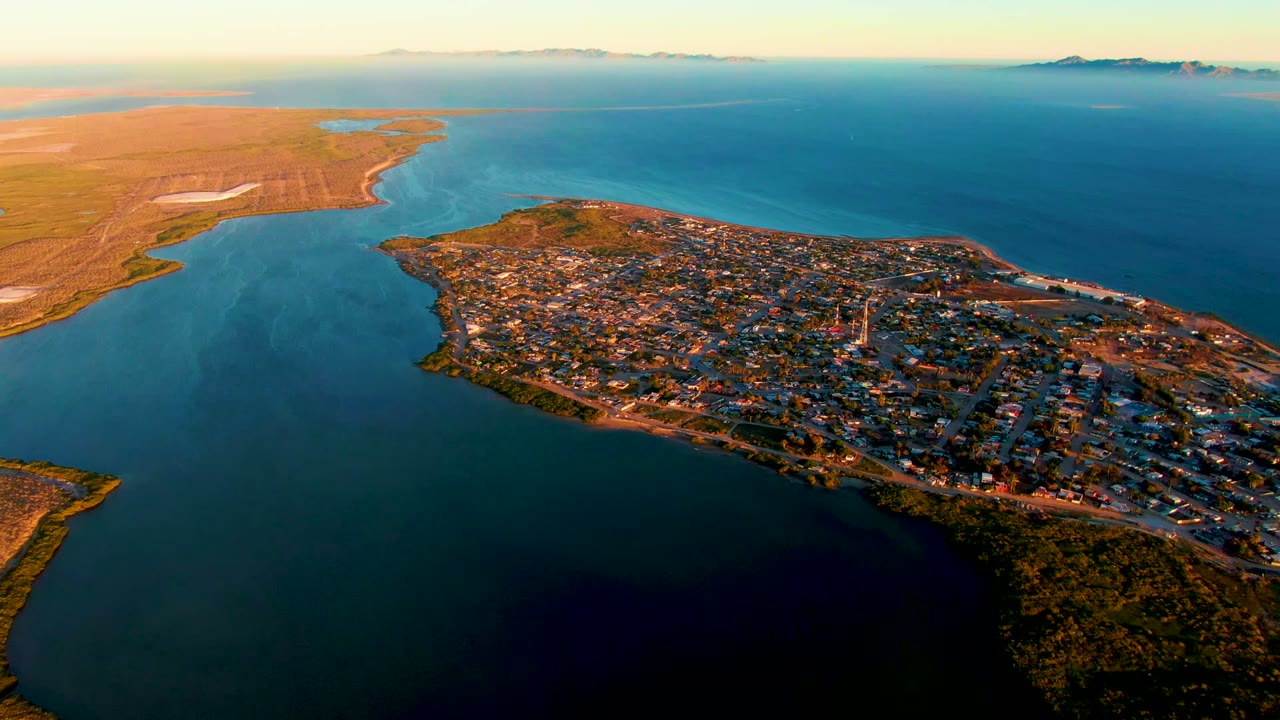 Opening aerial frame over San Carlos, Baja California Sur, showing the peninsula town bordered by dark foreground water, lighter ripples to the left, distant horizon, and no visible people.