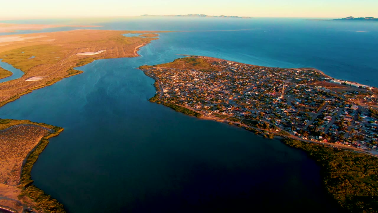 High aerial view over San Carlos, Baja California Sur, showing the coastal town, curving shoreline, blue-green bay, tan flats, distant horizon, and no visible people.