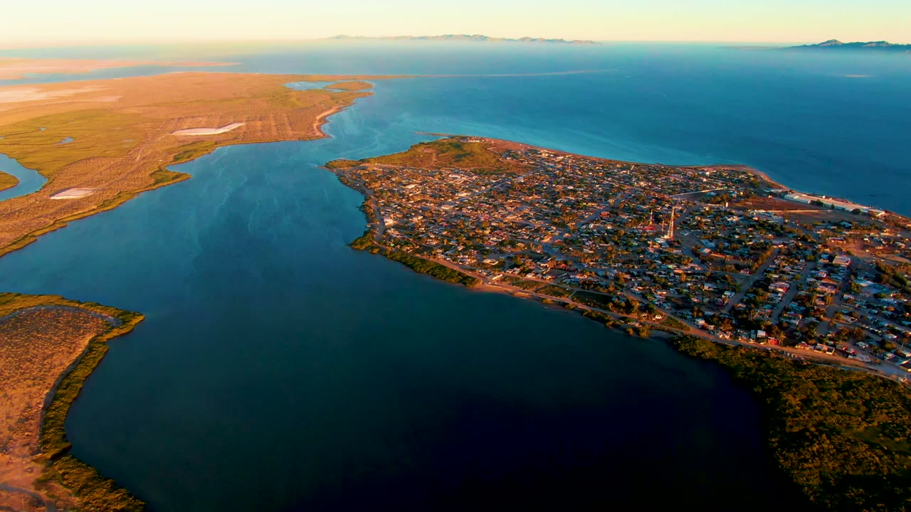 Wide drone view of San Carlos, Baja California Sur, with dense low buildings along the shore, open blue bay water below, tan coastal land beyond, and no visible people.