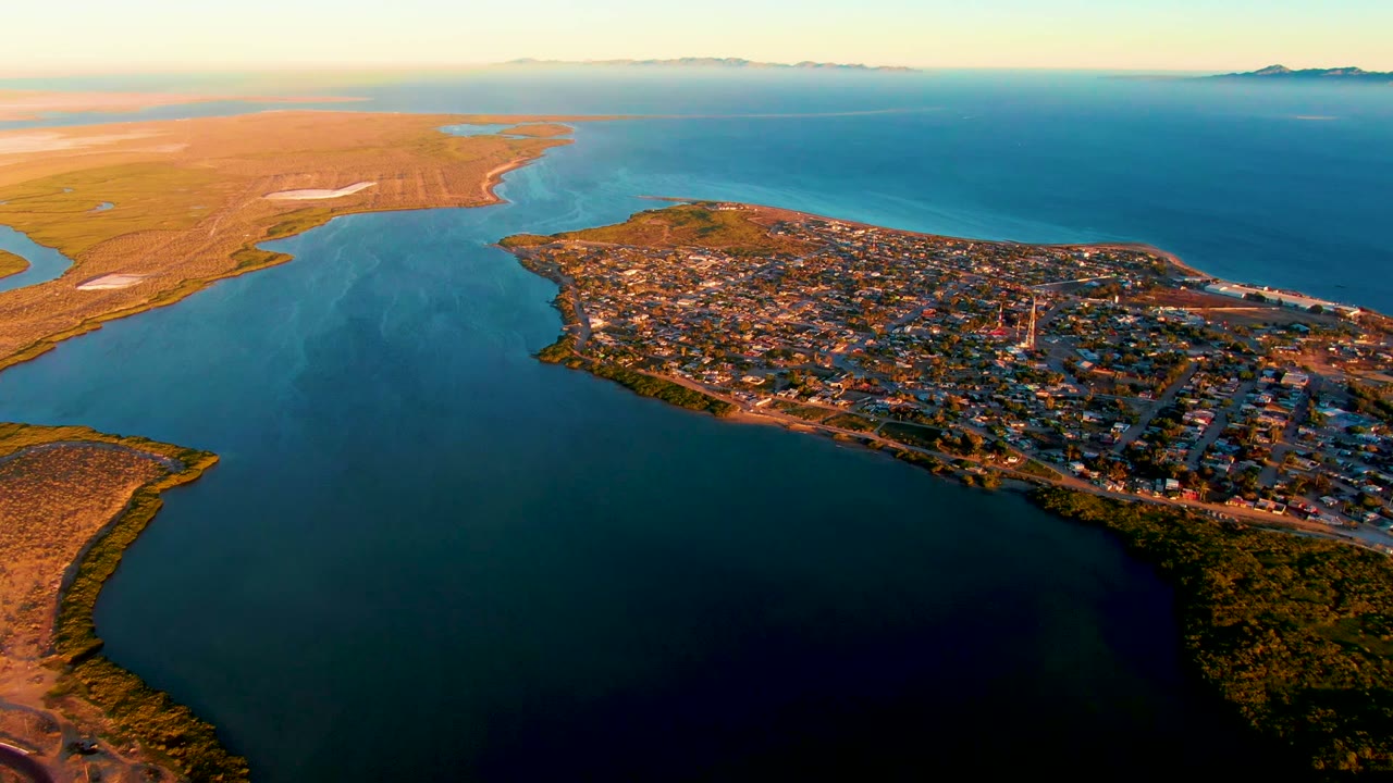 A high aerial frame over San Carlos, Baja California Sur shows dark bay water in front, a compact town on the right shoreline, golden grassy land to the left, distant horizon islands, and no visible people.