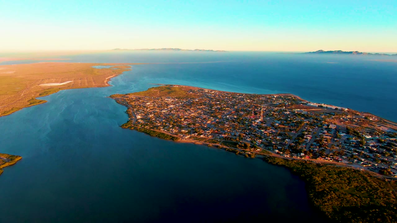Early frame of San Carlos, Baja California Sur, showing the town on a peninsula, blue inlet water in front, open land to the left, distant horizon, and no visible people.