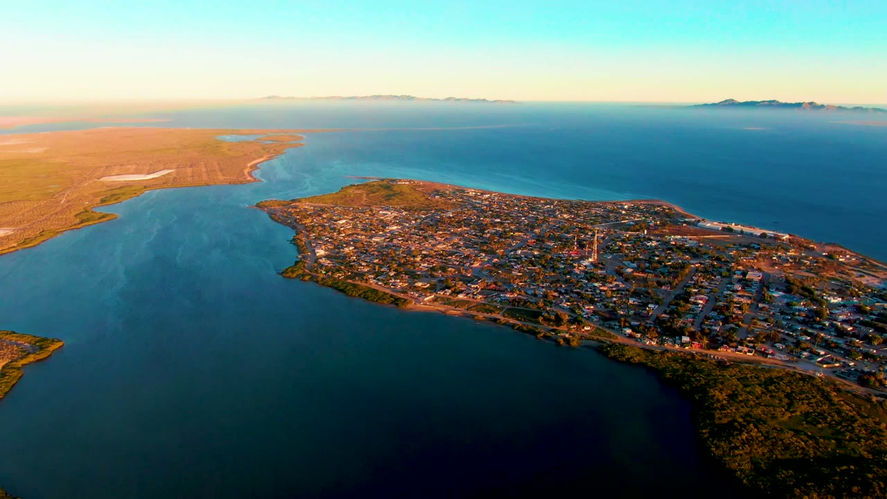 Aerial frame of San Carlos, Baja California Sur, with the peninsula town filling the lower right, calm inlet water across the foreground, ocean on the horizon, and no visible people.