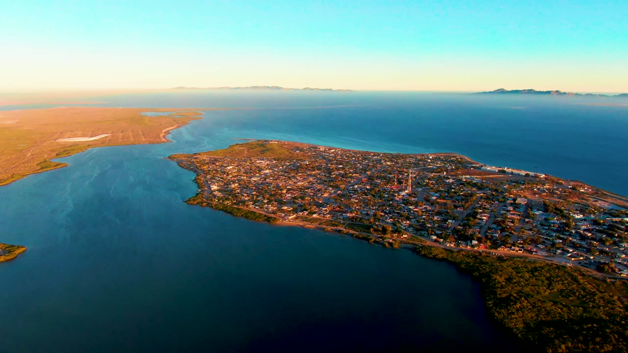 High aerial view over San Carlos, Baja California Sur, with the town on a peninsula, blue water on both sides, green vegetation at the lower edge, tan land beyond the shore, and no visible people.