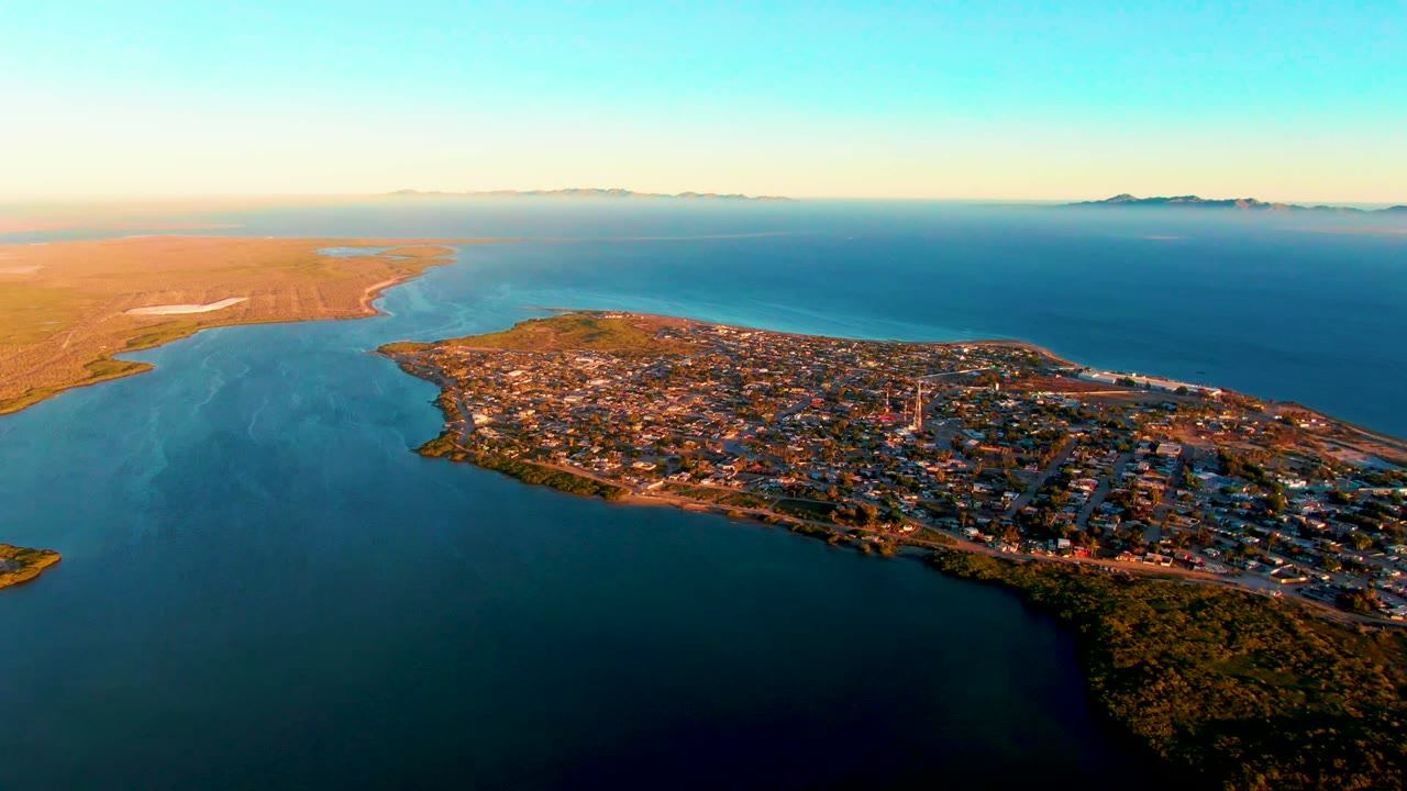 Static aerial over a coastal settlement with shoreline, inlet water, and distant hills.