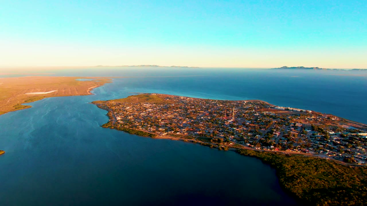 Aerial frame over San Carlos, Baja California Sur, showing a town on a peninsula with blue water in front, sandy land to the left, distant islands on the horizon, and no visible people.