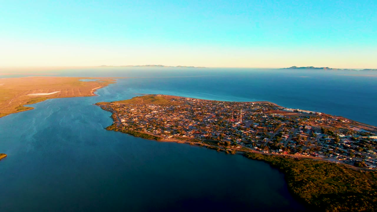 Wide drone view of buildings, shoreline, and open water under warm light.