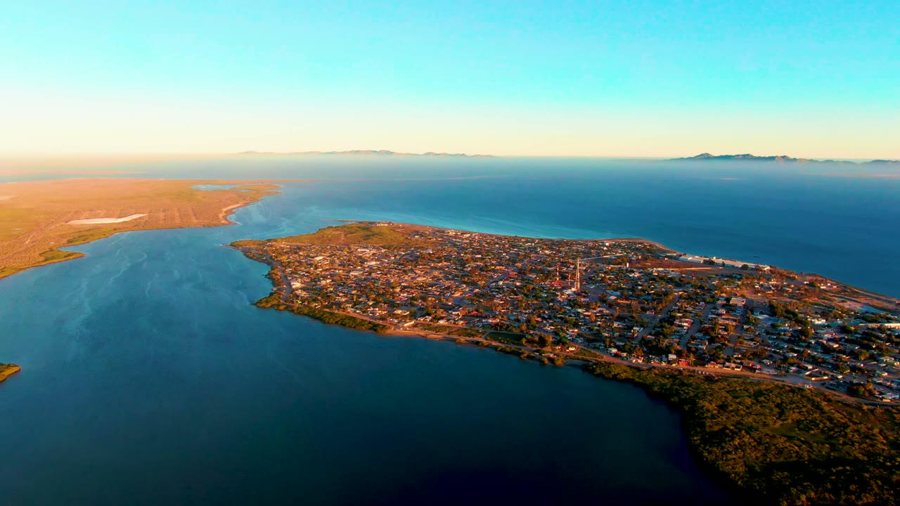 High aerial frame of San Carlos, Baja California Sur, with the town stretching along the shoreline, darker water in the foreground, pale land to the left, distant islands, and no visible people.