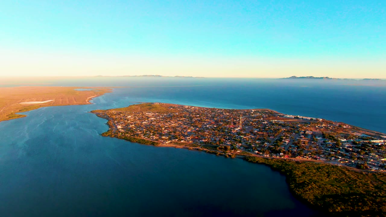 Wide aerial view over San Carlos, Baja California Sur, with a town on a peninsula between blue water and dry land, a bright land reflection at left, clear sky, and no visible people.
