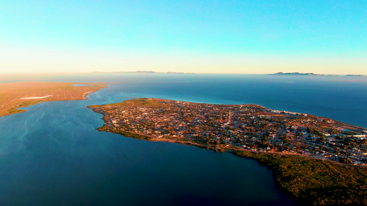 High-angle aerial over San Carlos, Baja California Sur, with the peninsula town stretching across the lower frame, open blue water to the right, sandy land to the left, and no visible people.