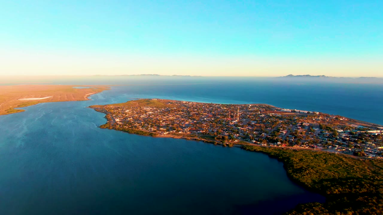 Aerial frame over San Carlos, Baja California Sur showing the peninsula town between blue water, sandy shoreline, distant horizon, warm dusk light, and no visible people.