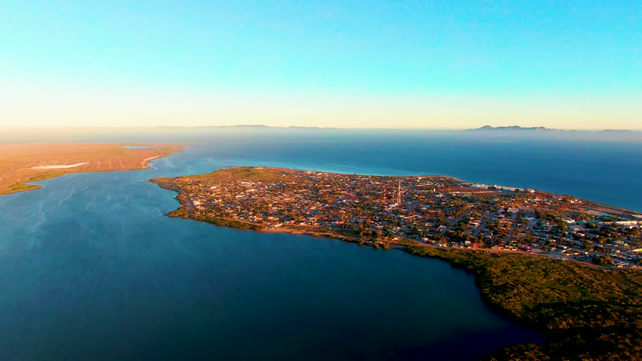 Wide drone view of peninsula buildings with ocean on both sides and a pale horizon.