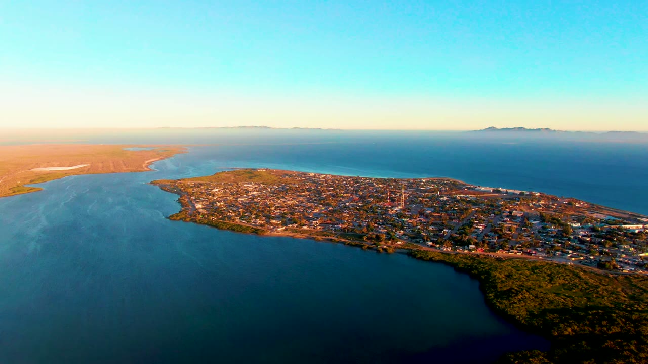 Aerial view over San Carlos at dusk with darker foreground water, sunlit town blocks, sandy coastline to the left, distant land on the horizon, and no visible people.