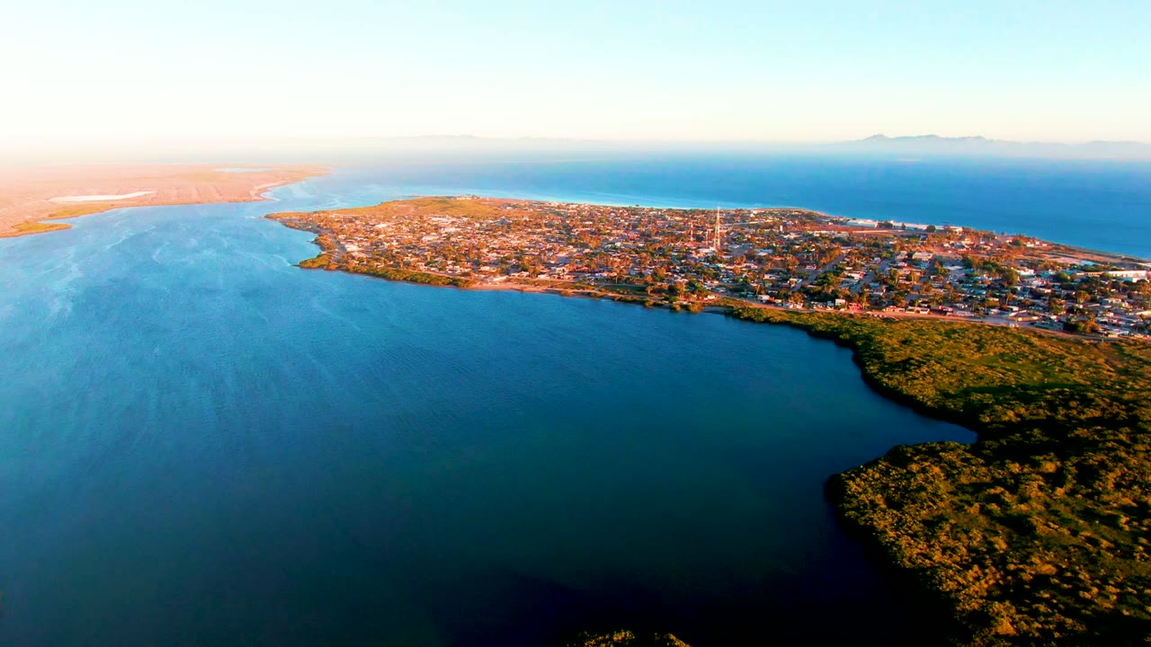 Aerial view over San Carlos, Baja California Sur, with a coastal town stretching across a narrow shoreline, blue water in the foreground and background, light mist on the left water, and no visible people.