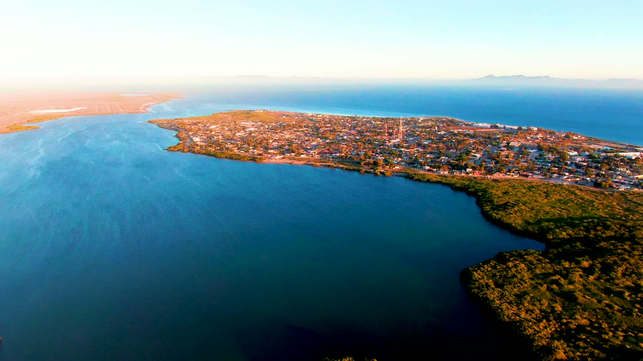 Wide drone view of a coastal settlement with calm water and haze near the shoreline.