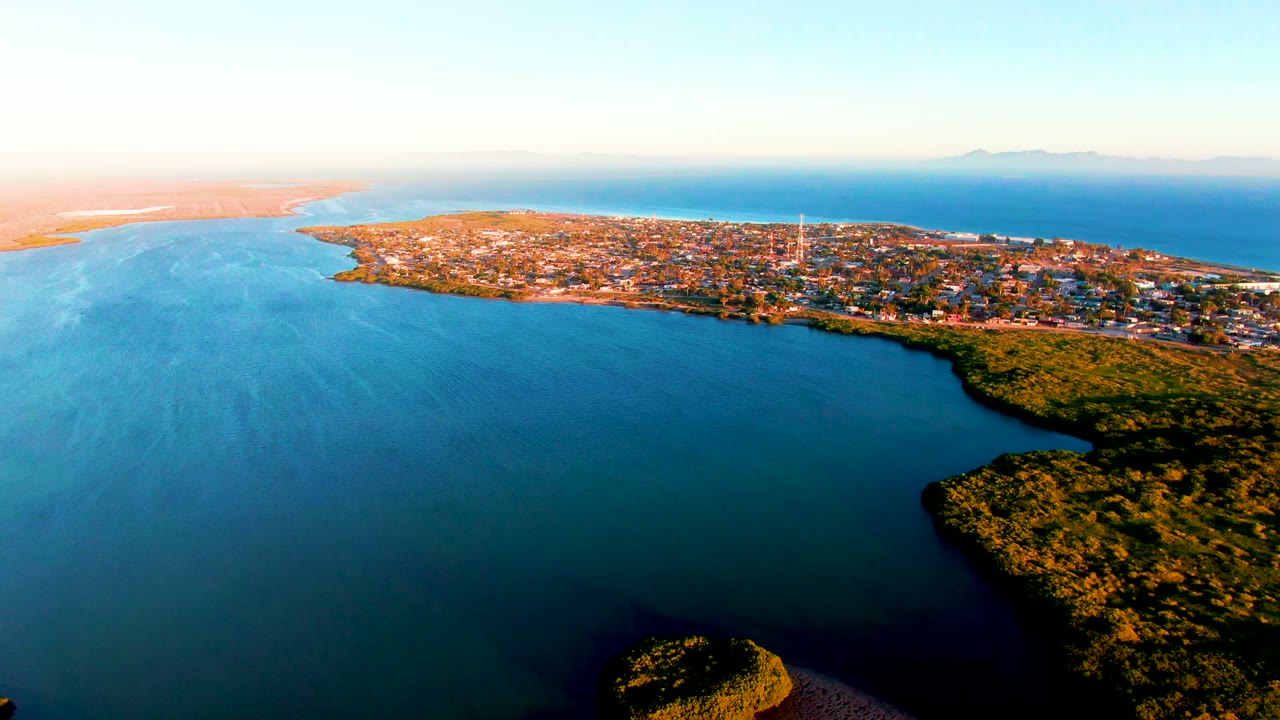 Aerial dusk view of San Carlos, Baja California Sur, with blue coastal water, green shoreline vegetation, a low peninsula town, distant horizon land, and no visible people.