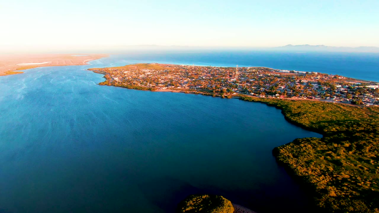 San Carlos, Baja California Sur appears across the midground under warm dusk light, with open blue water below, green coastal vegetation at right, distant landforms, and no visible people.