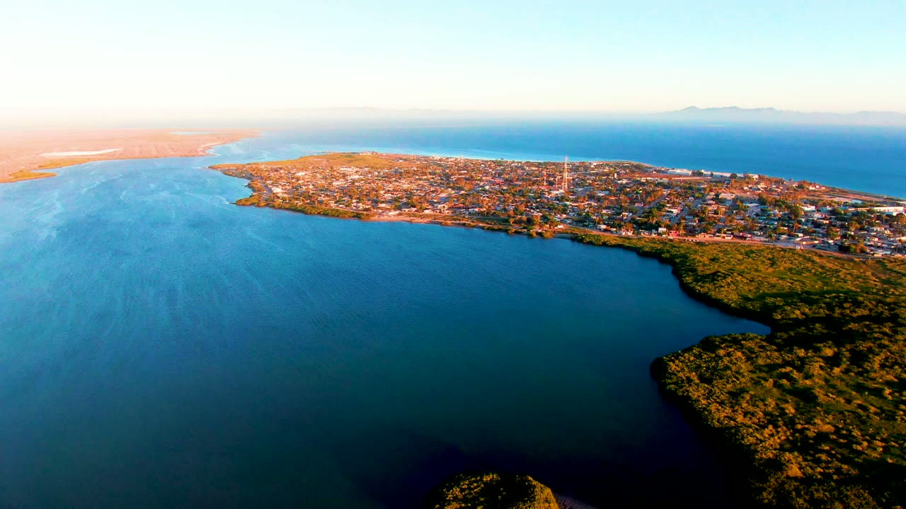 Aerial dusk view across calm water toward a low coastal town.