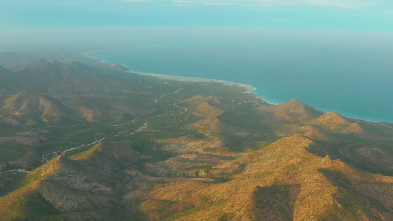 A high aerial frame over Cabo Pulmo, Baja California Sur shows sunlit brown hills, a winding road, sandy shoreline, blue ocean, hazy sky, and no visible people.