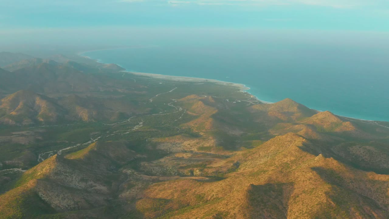 Aerial coastline with rocky hills, inland road lines, beach, and ocean horizon.