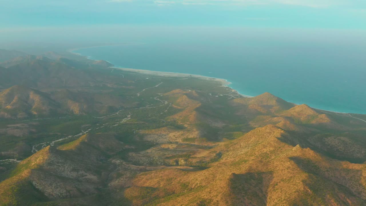 At two seconds, a wide drone view over Cabo Pulmo shows shadowed mountain slopes, golden hilltops, a pale shoreline, turquoise water, and no visible people.