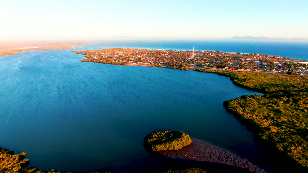 Aerial frame over San Carlos, Baja California Sur, with blue bay water in the foreground, a green shoreline at right, a low town across the coast, and no visible people.