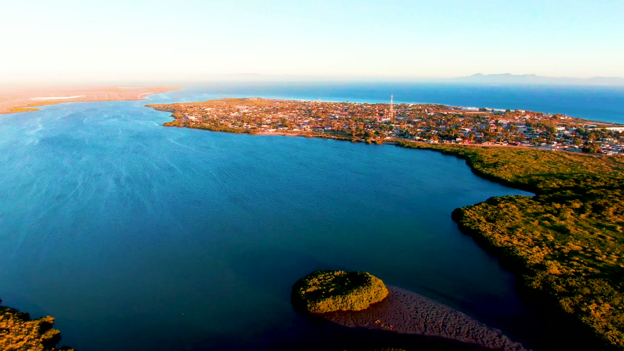 Wide aerial over bay water with town, shore vegetation, and warm light.