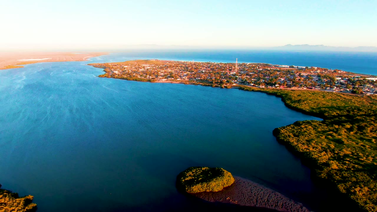 Drone-like aerial over San Carlos, Baja California Sur, with broad blue bay water, a small rounded landform near the lower right edge, shoreline greenery, town buildings, and no visible people.