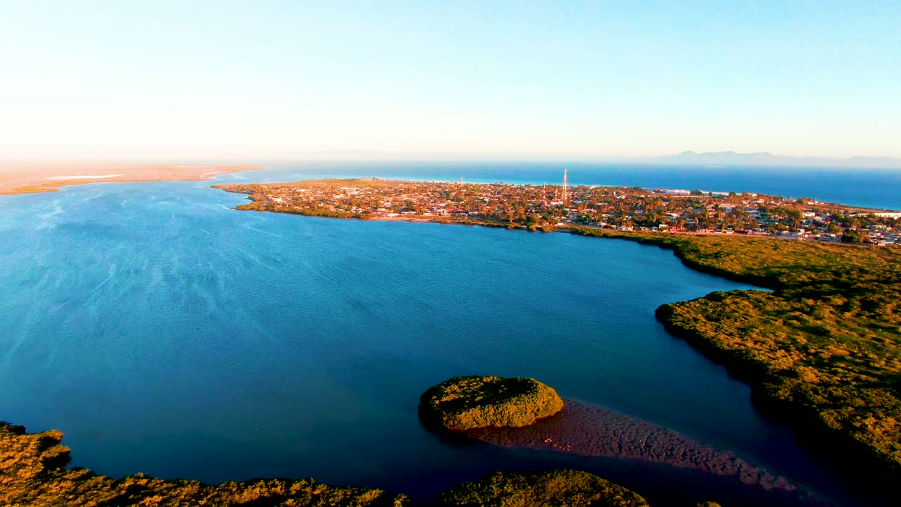 Aerial sunrise view over San Carlos, Baja California Sur, with calm blue water, a small green island low in frame, shoreline vegetation, a distant town, and no visible people.