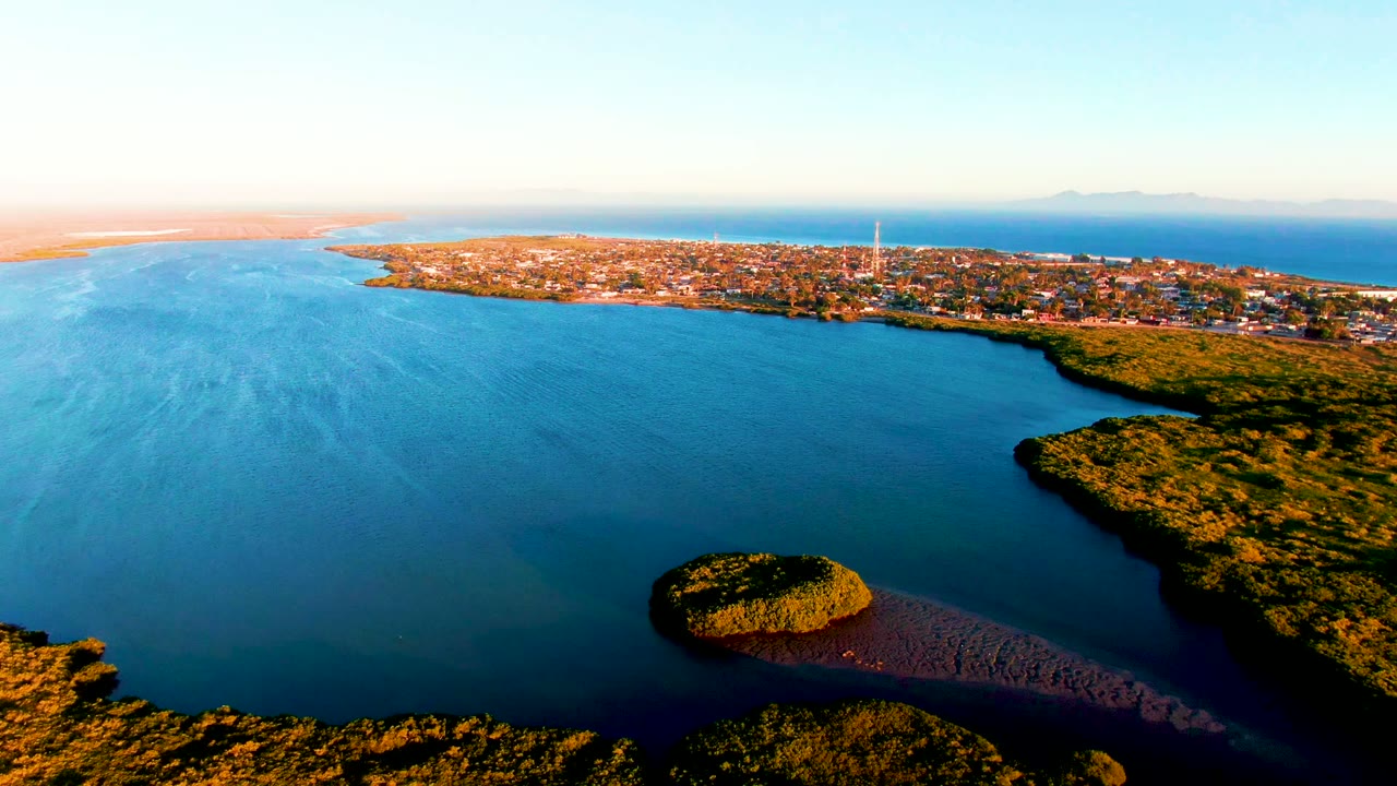 Wide aerial frame over San Carlos, Baja California Sur, showing the small island slightly right of center below the bay, a green shoreline edge, the town across the water, and no visible people.