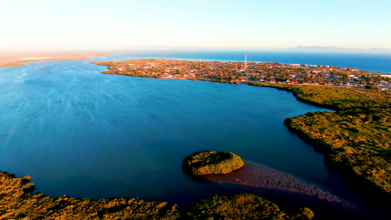 Aerial view in San Carlos, Baja California Sur, with blue water filling most of the frame, the small island low near center, dense green shoreline at right, distant town at top, and no visible people.