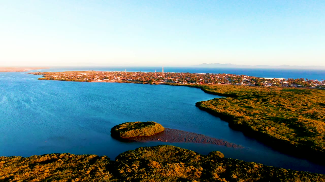 Wide aerial over a blue bay with a small island, green shoreline, coastal buildings, and clear sky.