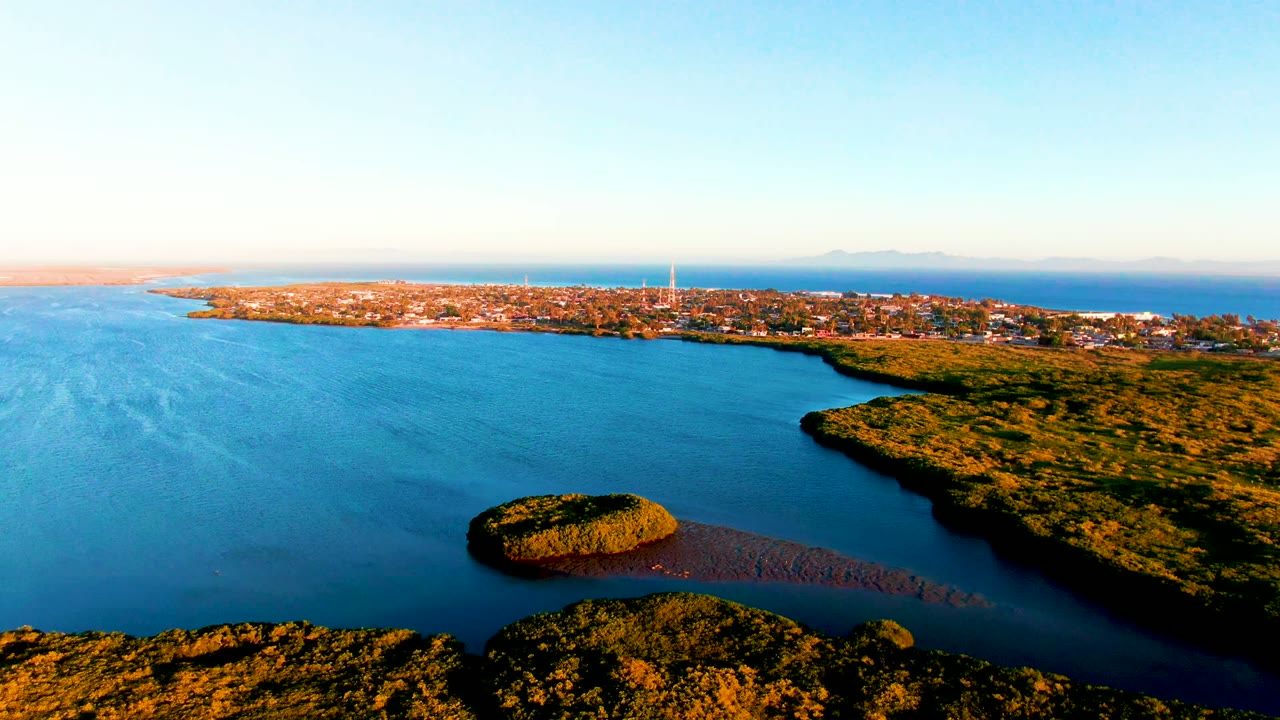 Aerial view of San Carlos, Baja California Sur with more open bay water visible on the left, the small green island below center, a darker shoreline edge to the right, distant town buildings, and no visible people.