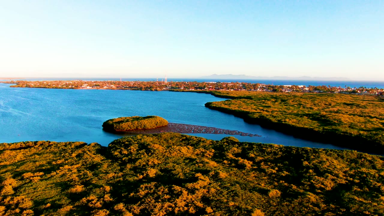 Opening aerial view of San Carlos, Baja California Sur, with green foreground vegetation, blue bay water, a small island-like patch near shore, distant town buildings, and no visible people.