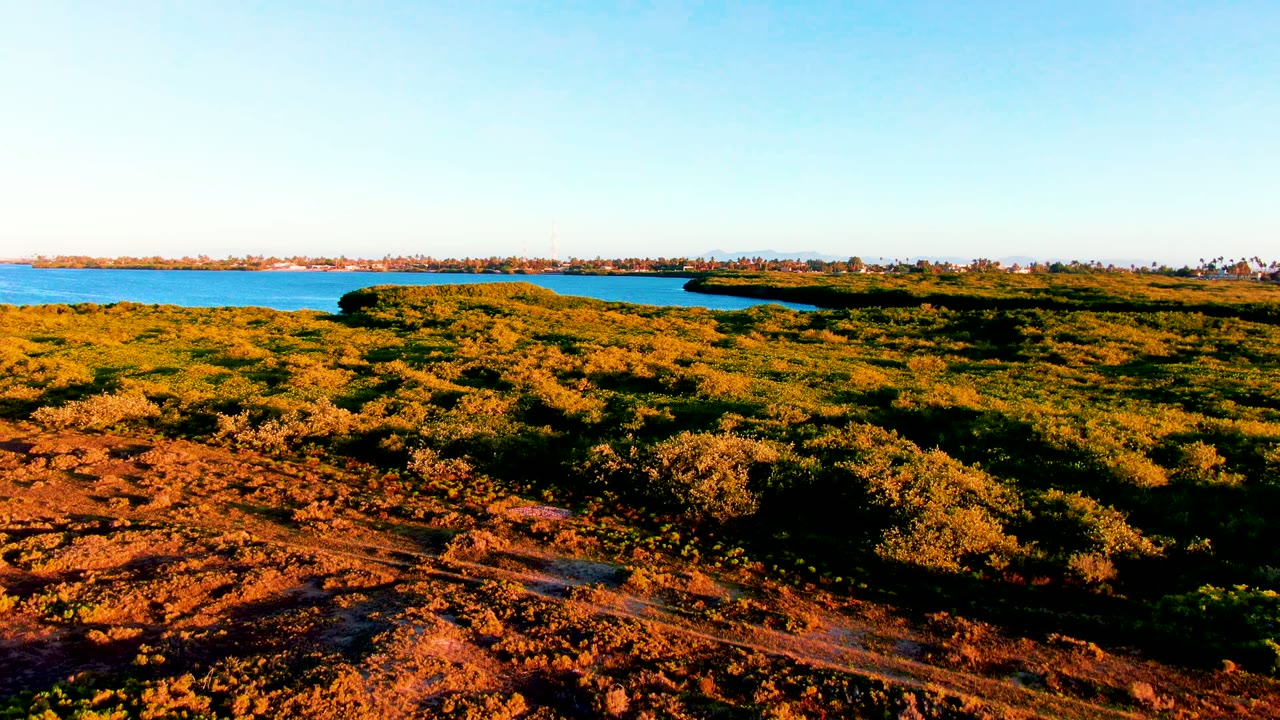 Opening aerial frame over San Carlos, Baja California Sur, with dirt and low green-gold vegetation in front, blue lake behind it, distant shoreline on the horizon, and no visible people.