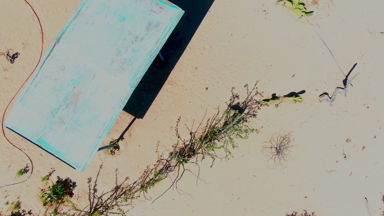 Overhead view in San Carlos, Baja California Sur of a weathered blue platform partly entering frame on pale sand, with sparse dry vegetation, a curved cable-like line, and no visible people.