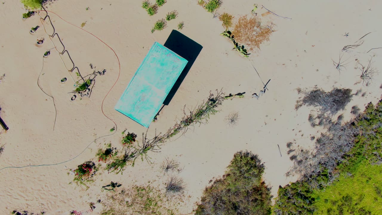 Overhead frame in San Carlos, Baja California Sur showing a turquoise rectangular structure near the upper left on pale sand, with scattered vegetation and no visible people.