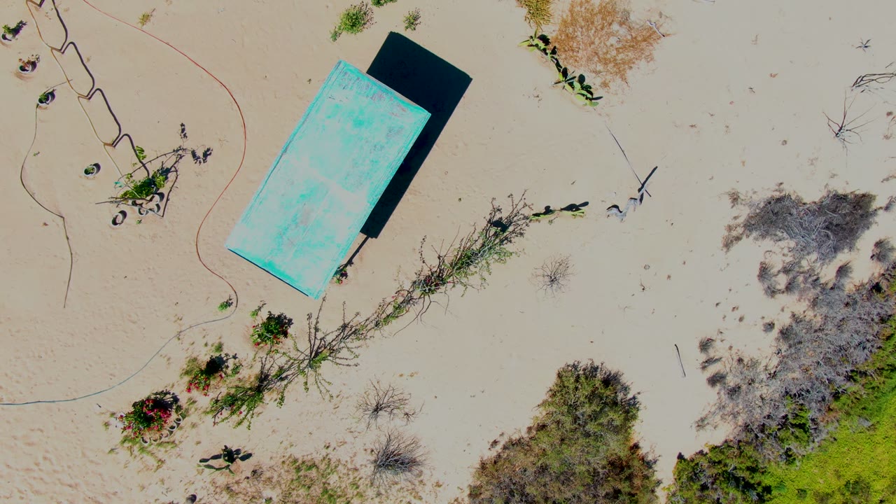 Overhead San Carlos frame where the turquoise structure sits toward the upper center, bordered by pale sand, low shrubs, dry brush, and no visible people.