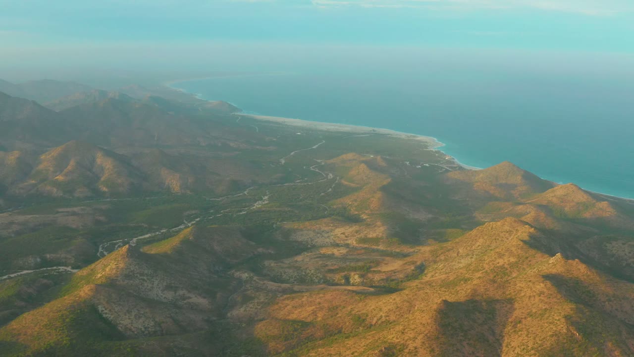 Aerial view over Cabo Pulmo coastal hills with a winding valley road, sandy shoreline, blue ocean, hazy horizon, and no visible people.