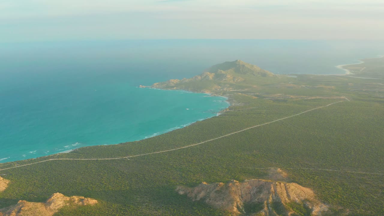 Opening aerial frame over Cabo Pulmo, Baja California Sur, with turquoise ocean, a curved shoreline road, green coastal vegetation, distant hills, and no visible people.