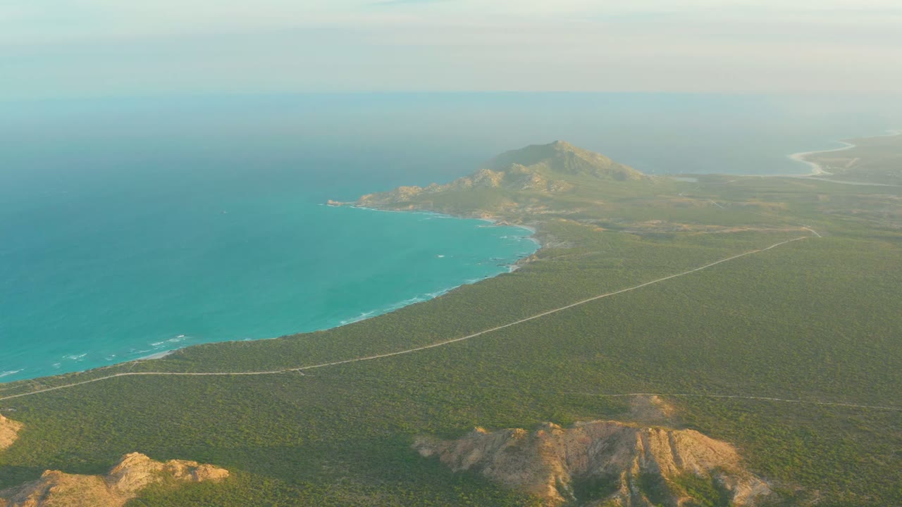Wide aerial coast with blue water, road, vegetation, and hills.